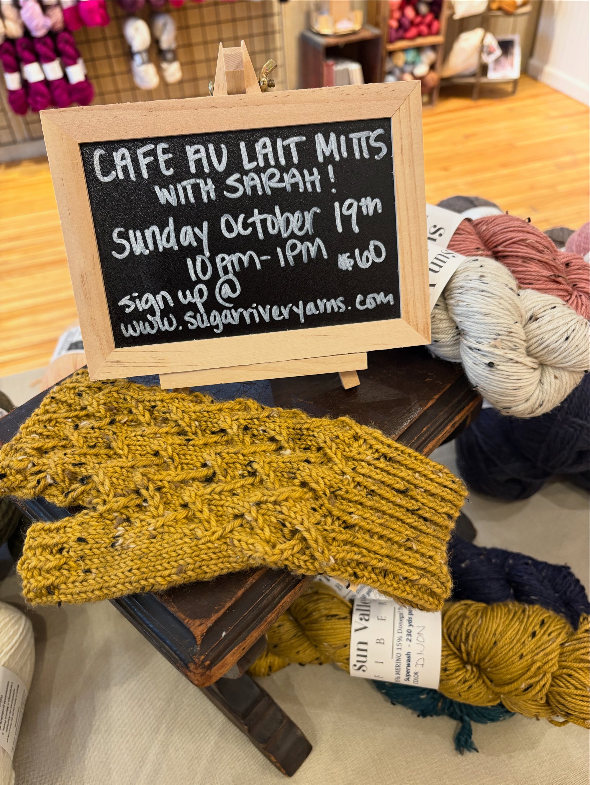 Knitted yellow mitten with a chalkboard sign advertising Cafe Au Lait Mitts class at Sugar River Yarns in Mt. Horeb, Wisconsin.