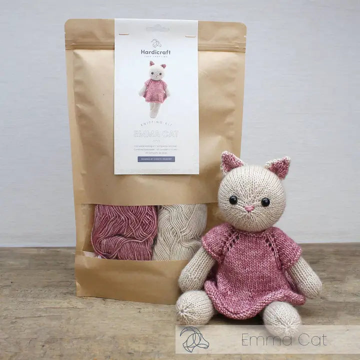 Knitted cat toy in a pink dress next to a package of yarn on a wooden surface.