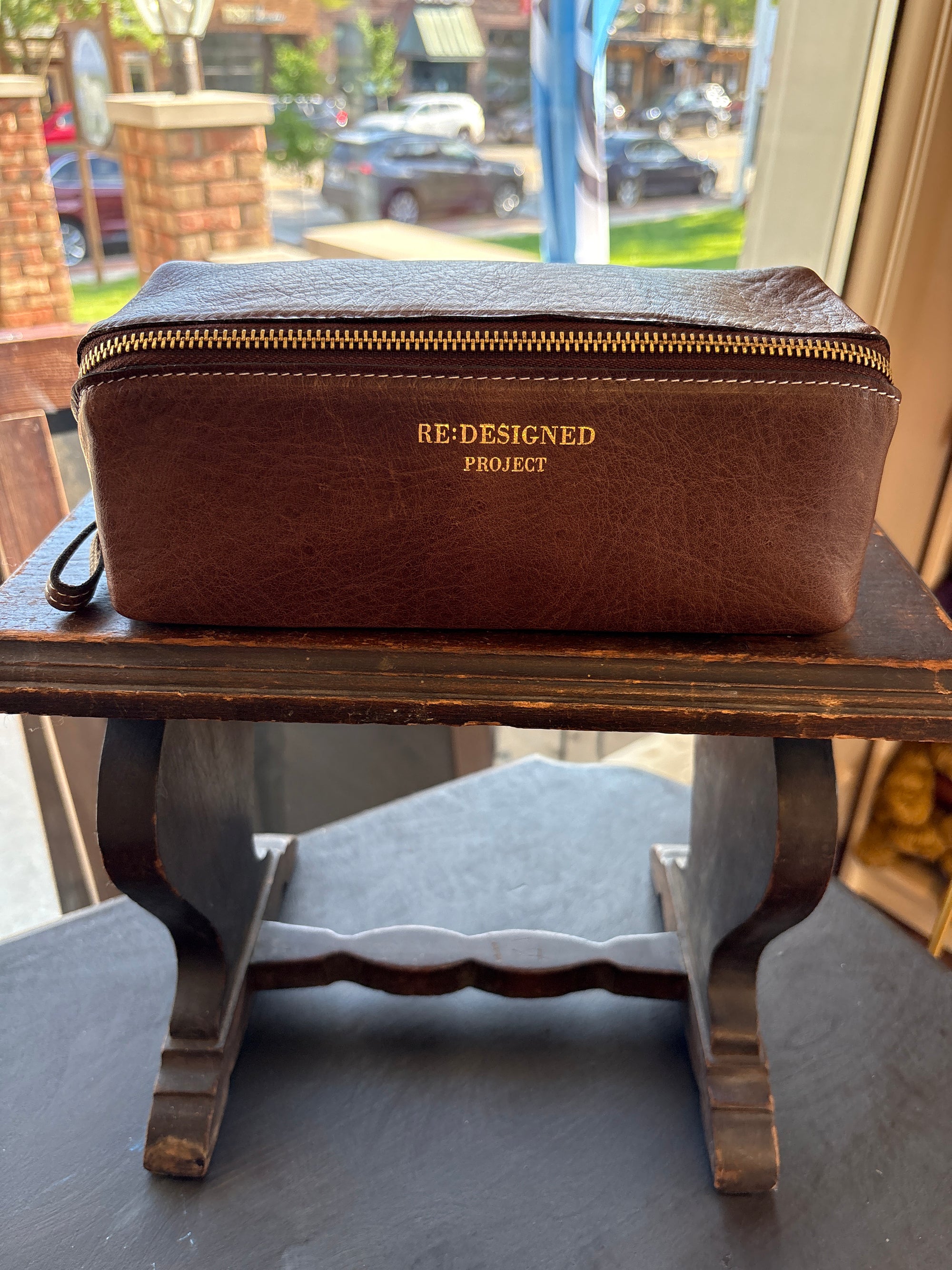 Brown leather knitting project bag with 'Redesigned Project' branding on a wooden stool.