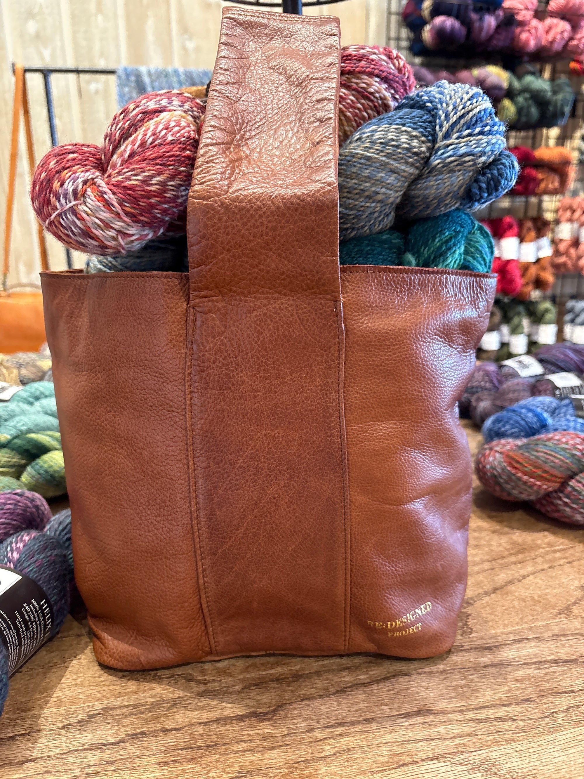 Project 81 brown leather bag filled with colorful yarn balls on a wooden surface.